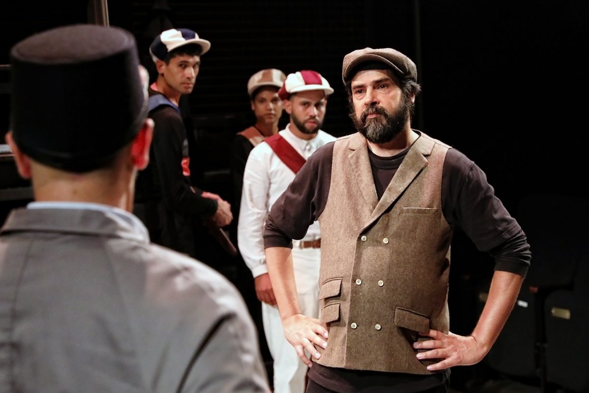 A bearded man in a brown waistcoat and cap stands with hands on hips, facing another man in a grey jacket and black hat, while four other men in uniforms and hats watch in the background on a dimly lit stage.