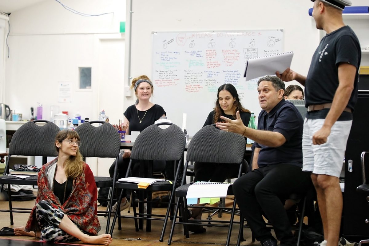 A group of people in a casual indoor setting talk and smile. One man stands holding papers; others sit in chairs or on the floor. A whiteboard with notes is visible in the background. The atmosphere appears collaborative and friendly.