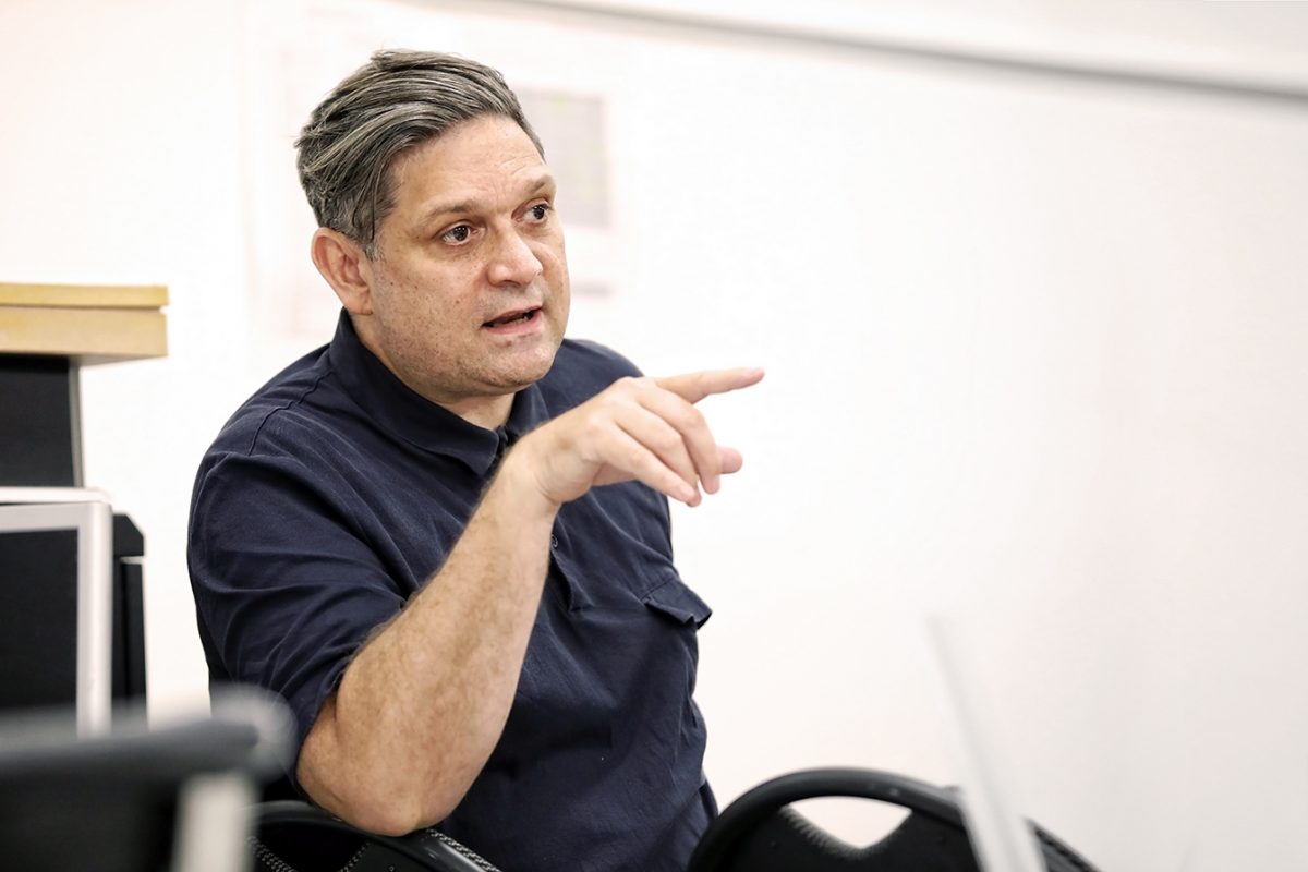 A man with short, dark hair streaked with grey, wearing a navy blue shirt, sits indoors and gestures with his hand while speaking, appearing engaged in conversation.