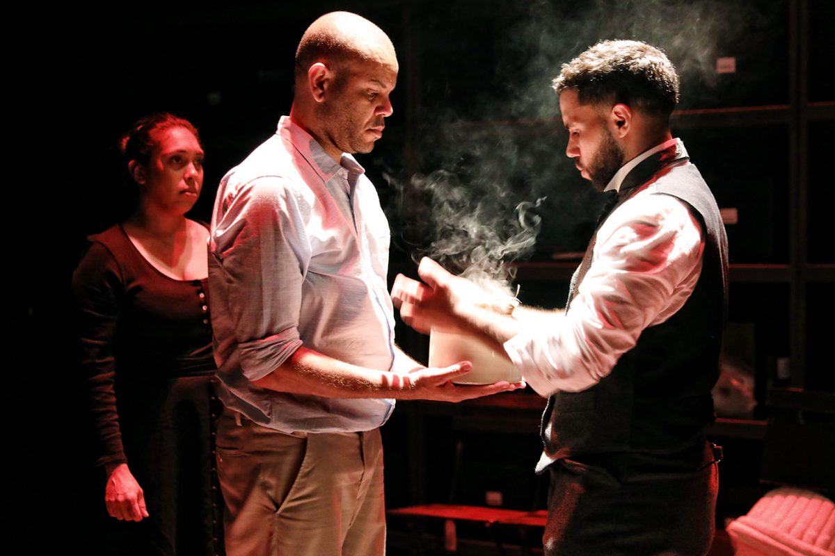Three people stand under dramatic lighting. One man holds a smoking box while another man receives it; a woman in dark clothing watches them with a serious expression. The background is dark and shadowy.