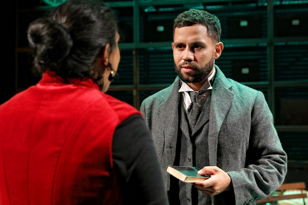 A man in a grey suit and overcoat holds a book while speaking to a woman in a red waistcoat. They appear to be on a dimly lit stage, suggesting a theatrical performance.