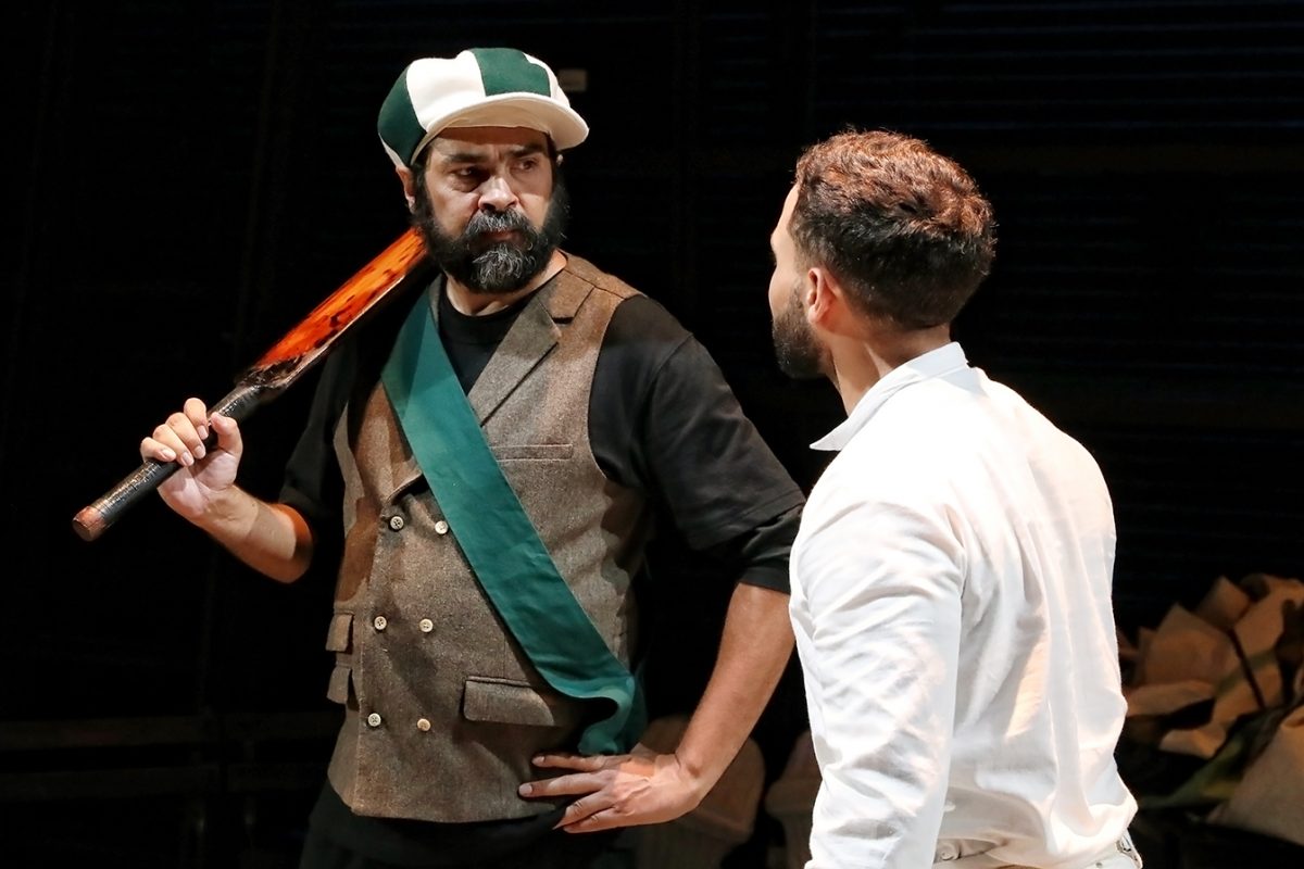 Two men face each other on stage; one wears a baseball cap, waistcoat, and green sash while holding a bat over his shoulder, looking serious. The other, dressed in white, stands with his back mostly to the camera.