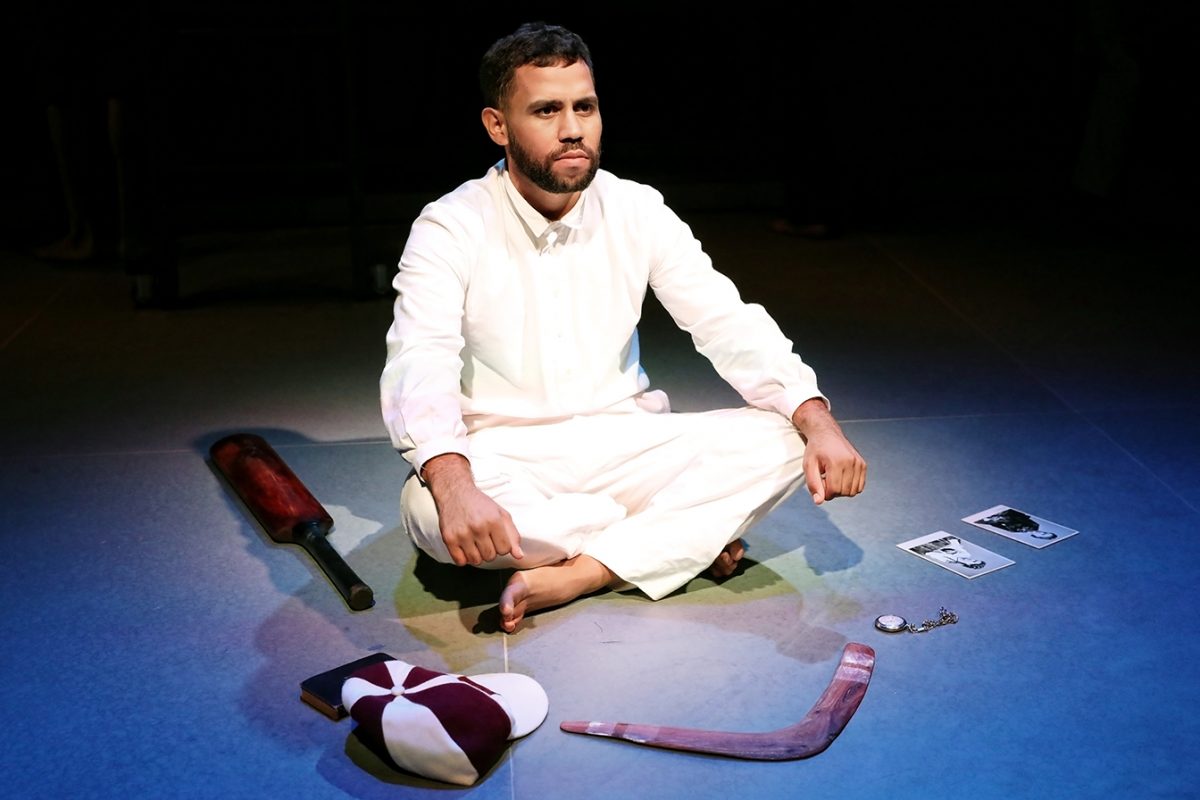 A man in a white outfit sits cross-legged on stage, surrounded by a cricket bat, cap, boomerang, photographs, and a pocket watch, under a spotlight.