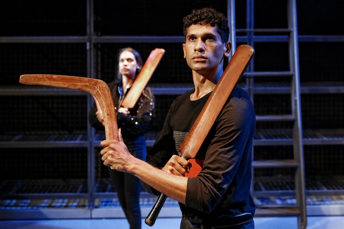 Two people stand holding wooden boomerangs in a dramatic pose on a stage with metal scaffolding in the background. The person in front looks determined, whilst the person behind looks focused. The lighting is theatrical.