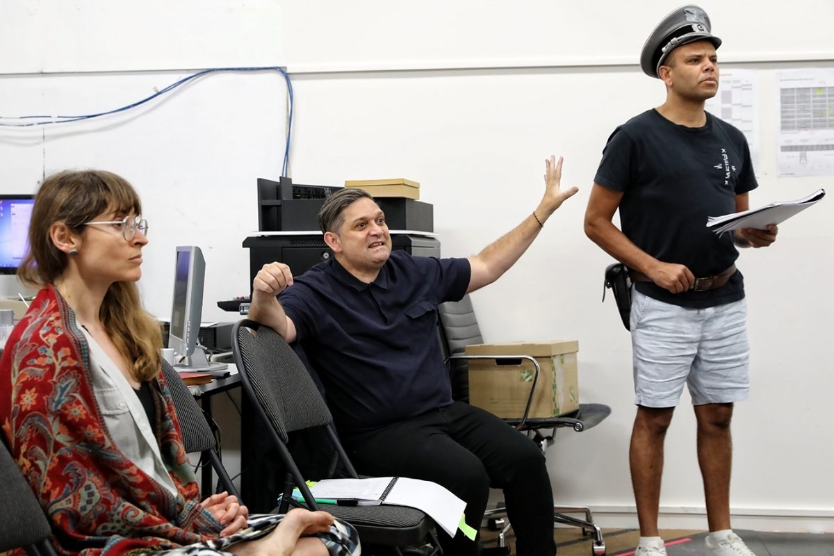 Three people indoors: a woman in glasses and a patterned blanket sits, a man in black gestures while seated, and another man stands wearing a police-style hat, dark T-shirt, and shorts, holding papers. Computer equipment is visible behind them.