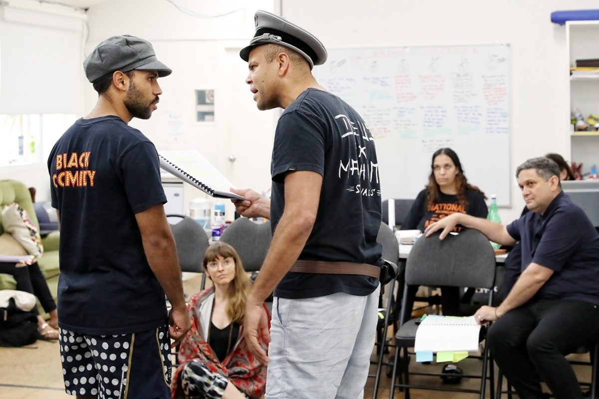 Two men in costume rehearse a dramatic scene, one confronting the other, whilst four people watch attentively in a classroom setting with scripts and a whiteboard in the background.