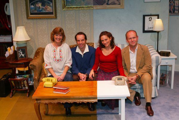 Four adults sit on a sofa and chairs in a cosy, warmly-lit living room set, surrounded by paintings, lamps, and vintage telephones on the tables in front of them. They are smiling and facing the camera.