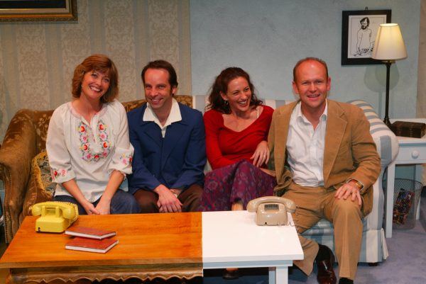 Four adults sit closely on a sofa, smiling at the camera. The group includes two women and two men. In front of them is a wooden table with two retro telephones and books. There is framed art and a lamp in the background.