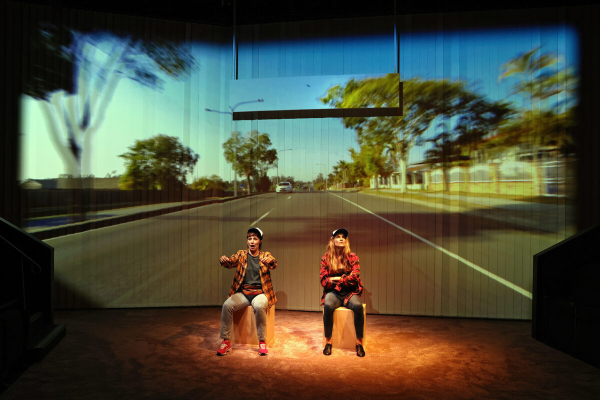 Two people sit on stage, miming driving with steering wheels, whilst a large projected image of a tree-lined road creates the illusion they are moving in a car. The scene is brightly lit and theatrical.
