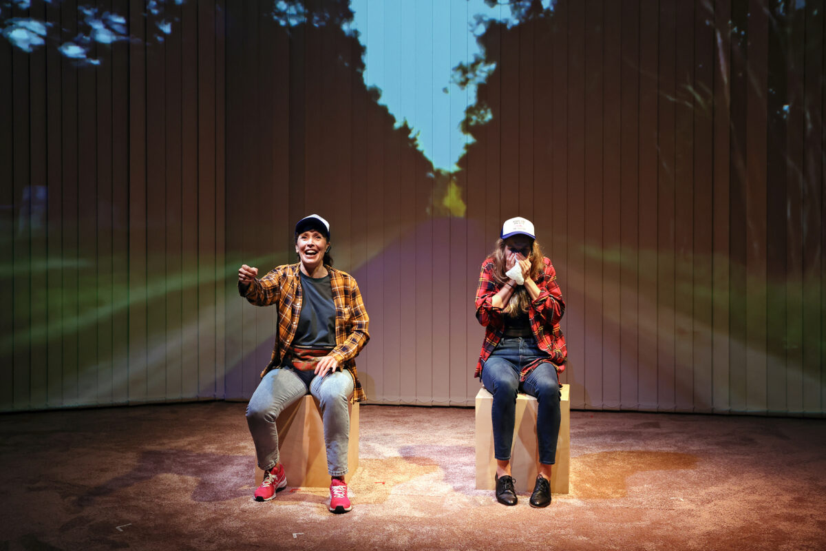 Two people in checked shirts and caps sit on stools on a stage, miming driving a car. A country road is projected behind them, creating the illusion they are travelling through the countryside.