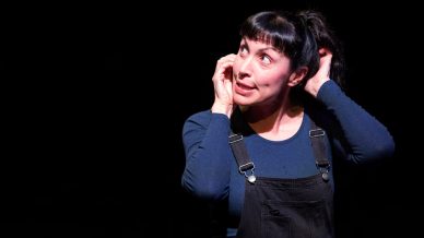 A woman with dark hair in a ponytail, wearing a navy long-sleeve shirt and black dungarees, stands against a black background, looking up and to the side while tugging at her ear with a curious expression.