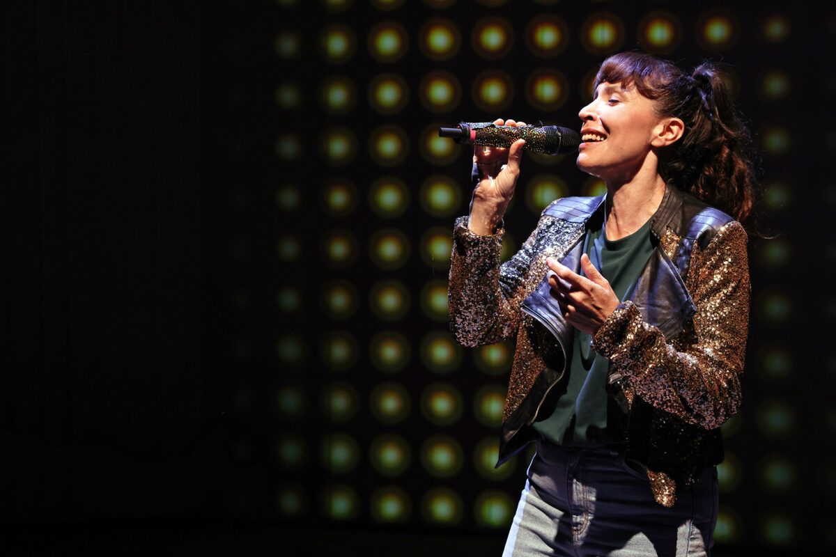 A woman wearing a sequinned jacket and jeans sings passionately into a microphone on stage, with a background of circular lights creating a dramatic effect.