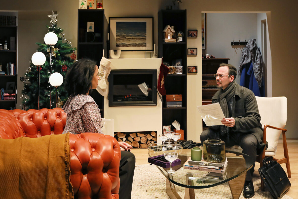 A woman sits on an orange sofa facing a man with glasses who holds papers in a cosy living room decorated for Christmas, with a tree, fireplace, and holiday stockings.