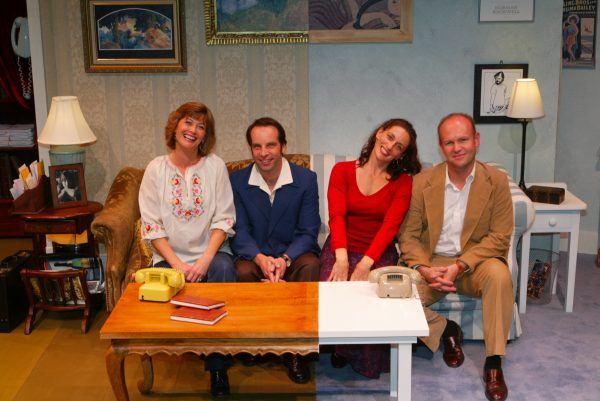 Four adults sit on a sofa in a lounge setting, smiling at the camera. Two women are at the ends, with two men in suits between them. The room is decorated with framed art, books, and lamps.