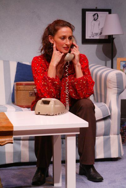 A woman with brown hair, wearing a red blouse and brown trousers, sits on a striped sofa and talks on a beige rotary telephone in a cosy living room setting. A suitcase and framed photo are visible in the background.