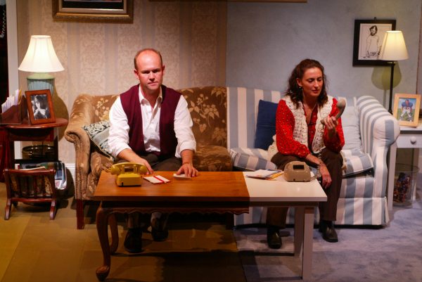 A man and a woman sit on separate sofas in a lounge, each with a rotary telephone on the coffee table between them. The man looks forward while the woman looks down, holding a telephone cord. The room is warmly lit and decorated.