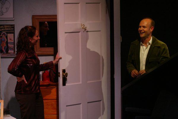 A woman stands by an open door marked 12F, smiling at a man standing outside. The room is dimly lit, with a chest of drawers, mirror, and a film poster on the wall. The man appears to be laughing.