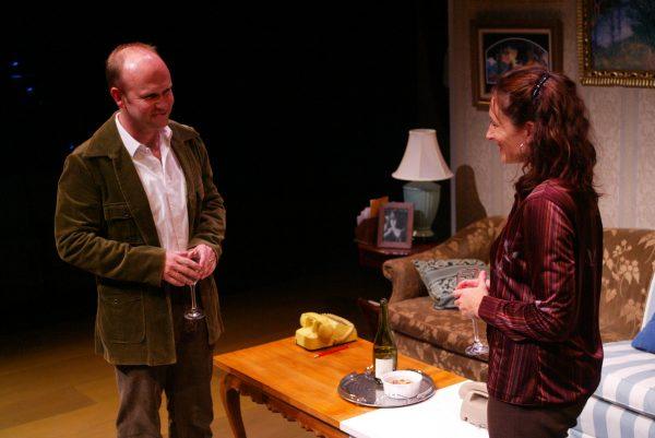 A man and a woman stand by a coffee table in a warmly lit sitting room, holding wine glasses and smiling at each other. The table has a wine bottle, glasses, and snacks. A sofa and framed pictures are in the background.