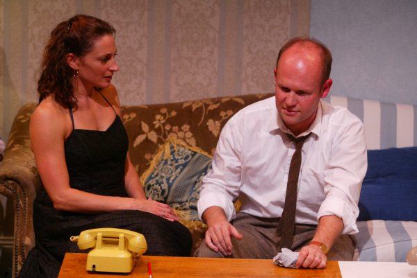 A woman in a black dress sits on a sofa beside a man in a white shirt and tie, who is seated at a wooden table with a yellow rotary telephone, a red pencil, and papers. Both appear deep in thought.