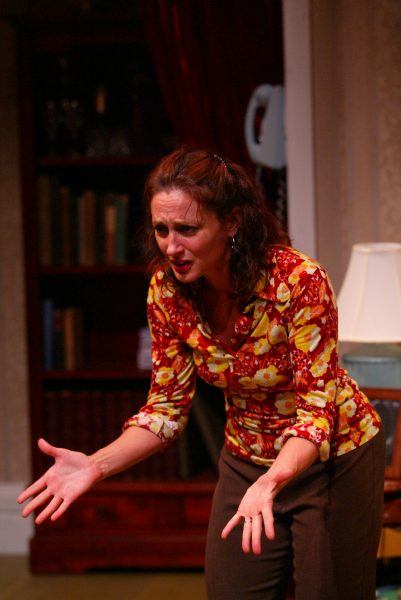 A woman with brown hair wearing a red and yellow floral blouse gestures emotionally with both hands, leaning forward in a room with shelves, books, and a lamp in the background.