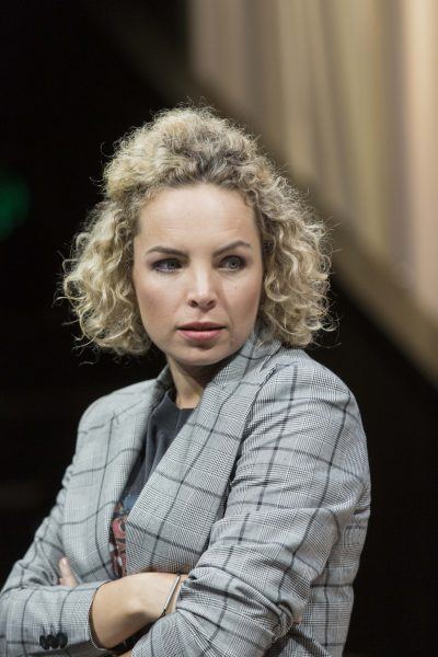 A woman with curly blonde hair wearing a grey checked blazer stands with her arms crossed, looking to the side with a serious expression. The background is softly blurred.