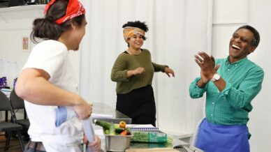 Three people in a kitchen laugh and interact. One person wears a red bandana, another has a colourful headwrap and green jumper, and the third, in a blue apron, claps happily. Fresh vegetables and utensils are on the counter.