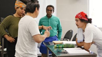 Four people have a serious discussion in a kitchen. One person gestures while talking, two listen attentively, and one leans on the counter beside papers, a tea towel, and a bunch of coriander. Cooking supplies are visible on the table.