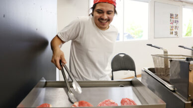 A person wearing a red bandana and white shirt smiles whilst cooking raw meat slices on a flat grill using metal tongs in a bright kitchen.