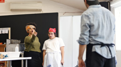 A woman gestures and points whilst speaking to two men in a kitchen setting; one man in a chef’s uniform looks surprised, and the other stands with his back to the camera.