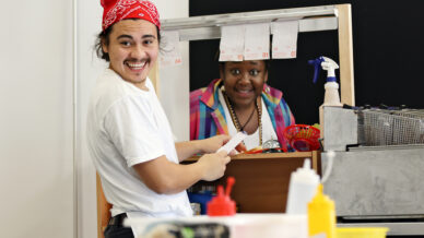 A smiling man wearing a red bandana holds a receipt by a food counter, whilst a woman in a colourful shirt smiles from behind the counter. Various condiments and food items are visible in the foreground.