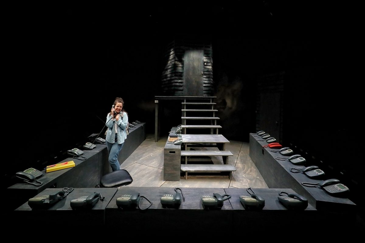A woman stands in a dimly lit room, speaking on a corded telephone. She is surrounded by many identical telephones arranged on a U-shaped desk, with stairs and a dark, industrial background behind her.