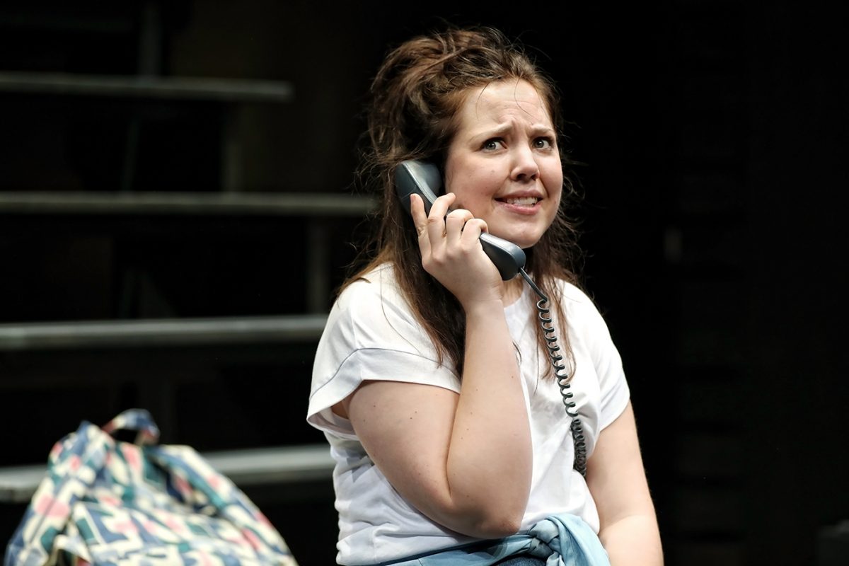 A woman with long brown hair, wearing a white T-shirt, holds a corded phone to her ear and has a concerned or worried expression. A patterned rucksack sits beside her on a bench, with a dark background behind.