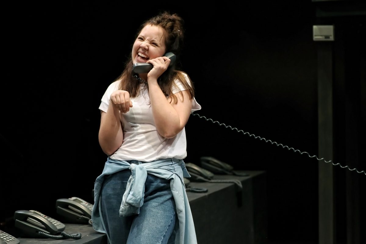 A young woman in a white T-shirt and jeans laughs while talking on a corded phone, with several other phones on the table beside her. She has a denim jacket tied around her waist and stands in a dark room.