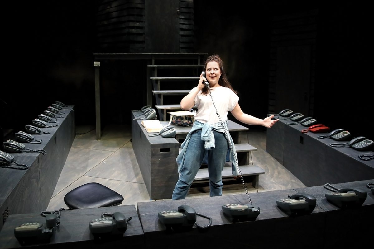 A woman stands in a modern stage set, holding a phone to her ear and smiling. She’s surrounded by a U-shaped desk lined with multiple black phones and a single red phone. The background is dark and minimalist.