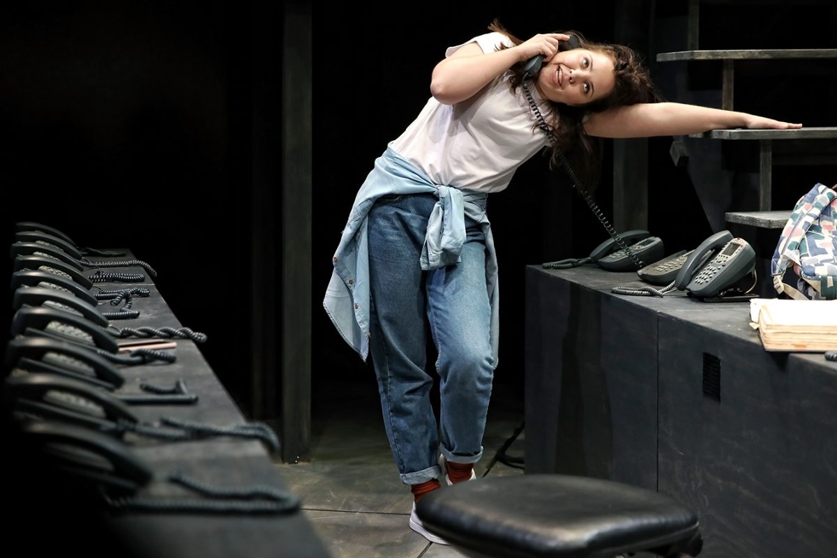 A young woman in jeans and a white shirt leans sideways while talking on a corded phone. She is in a room with a row of phones on a desk, and another phone is on the surface beside her.
