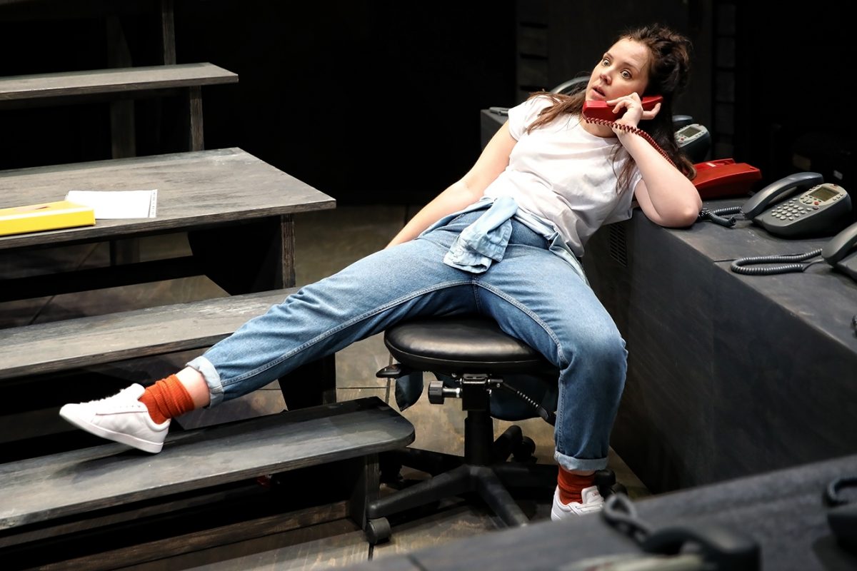 A woman in a white shirt and blue jeans lounges on a chair with her legs stretched out, talking on a red corded phone. She appears relaxed and casual in an office-like setting with multiple telephones on the desk.