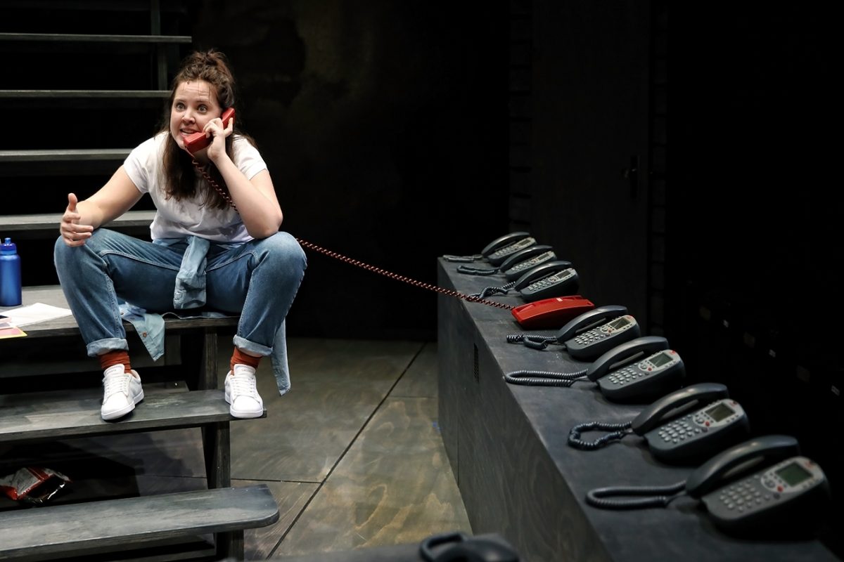 A woman in casual clothes sits on stairs, talking on a red corded phone. She gives a thumbs-up and is surrounded by several black telephones lined up on a platform beside her. The setting appears to be a stage.
