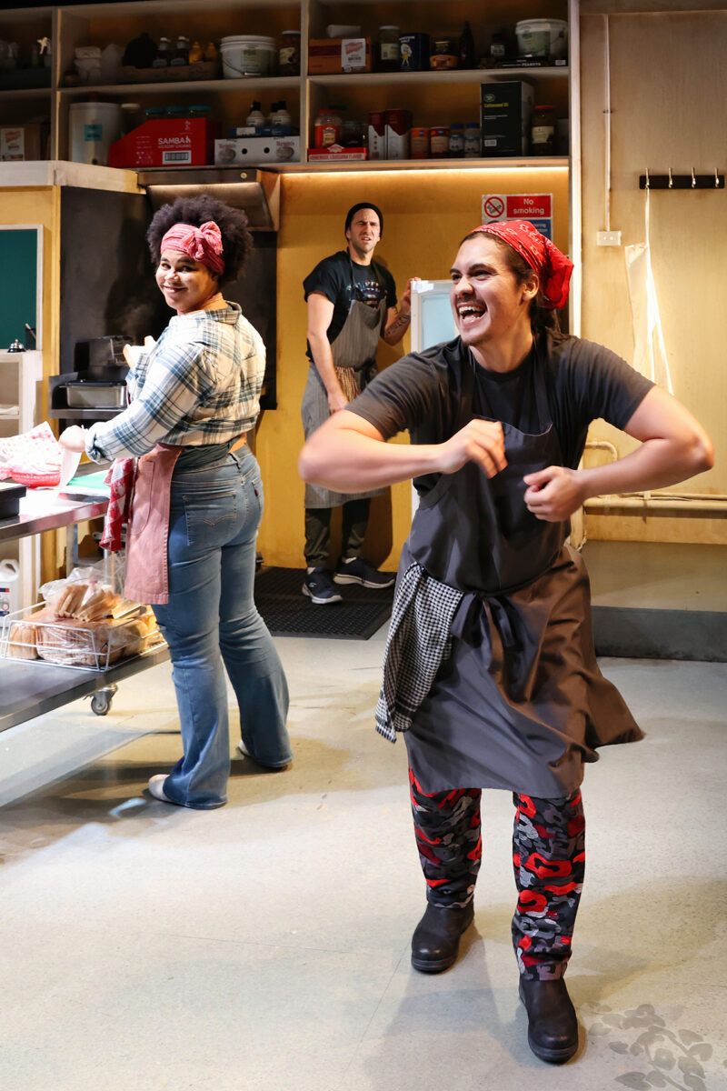 Three people are in a bakery kitchen. Two women in aprons and colourful headscarves are at the front, one smiling and the other dancing excitedly. A man stands in the background, watching and smiling. Bread and kitchen items are visible.