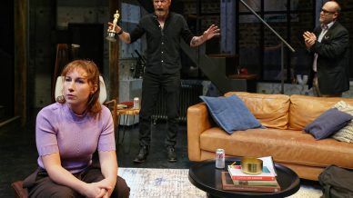A woman in a purple top sits on a sofa looking away, while a man in black stands behind her holding an Oscar trophy, and another man in a suit stands clapping near a staircase in a modern living room set.