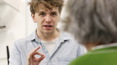 A young man with tousled hair wearing a light blue shirt gestures with his hand whilst speaking to an older person in a green top, who is seen from behind. The setting appears to be indoors.