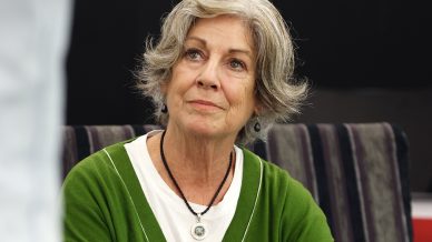 An older woman with grey hair and a green cardigan sits on a striped sofa, looking up thoughtfully at someone out of frame. A brown glass bottle and some papers are on the table in front of her.
