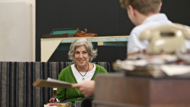 An older woman with grey hair and a green jumper smiles while seated opposite a younger person in a light shirt, who is holding papers. A vintage telephone, books, and papers are on the desk in the foreground.