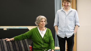 A woman with grey hair wearing a green cardigan sits and smiles gently whilst a young man in a light blue shirt stands beside her, laughing. They appear to be in a casual, indoor setting.