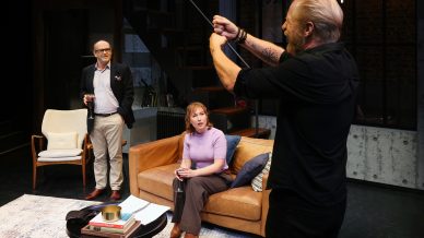 Three people are in a modern living room. A man in black stands holding a snooker cue, gesturing energetically. A seated woman and a standing man with glasses watch him, both holding wine glasses. The setting is cosy and warmly lit.