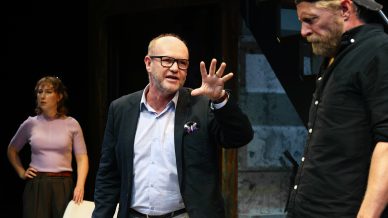 A man in a blazer gestures with his hand whilst speaking to another man in a black shirt and cap; a woman stands in the background watching the interaction.