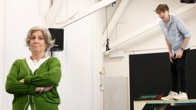 An older woman stands with arms folded in the foreground, looking ahead. In the background, a young man in a light shirt stands on a table, leaning forward and looking towards her in a bright room with exposed beams.
