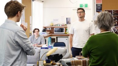Four people are in a casual office or classroom setting. One man in a white shirt stands and speaks, while others listen. The room has a sofa, desk, papers, and a microwave in the background.