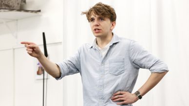 A young man with tousled hair and a light blue button-up shirt stands indoors, looking serious. He points forward with one hand while resting his other hand on his hip, against a white curtain background.