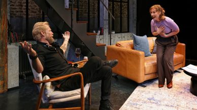 A man sits in an armchair with arms outstretched, looking at a woman who is standing, holding her chest and appearing emotional. They are in a modern lounge with a staircase, sofa, and cosy decor.