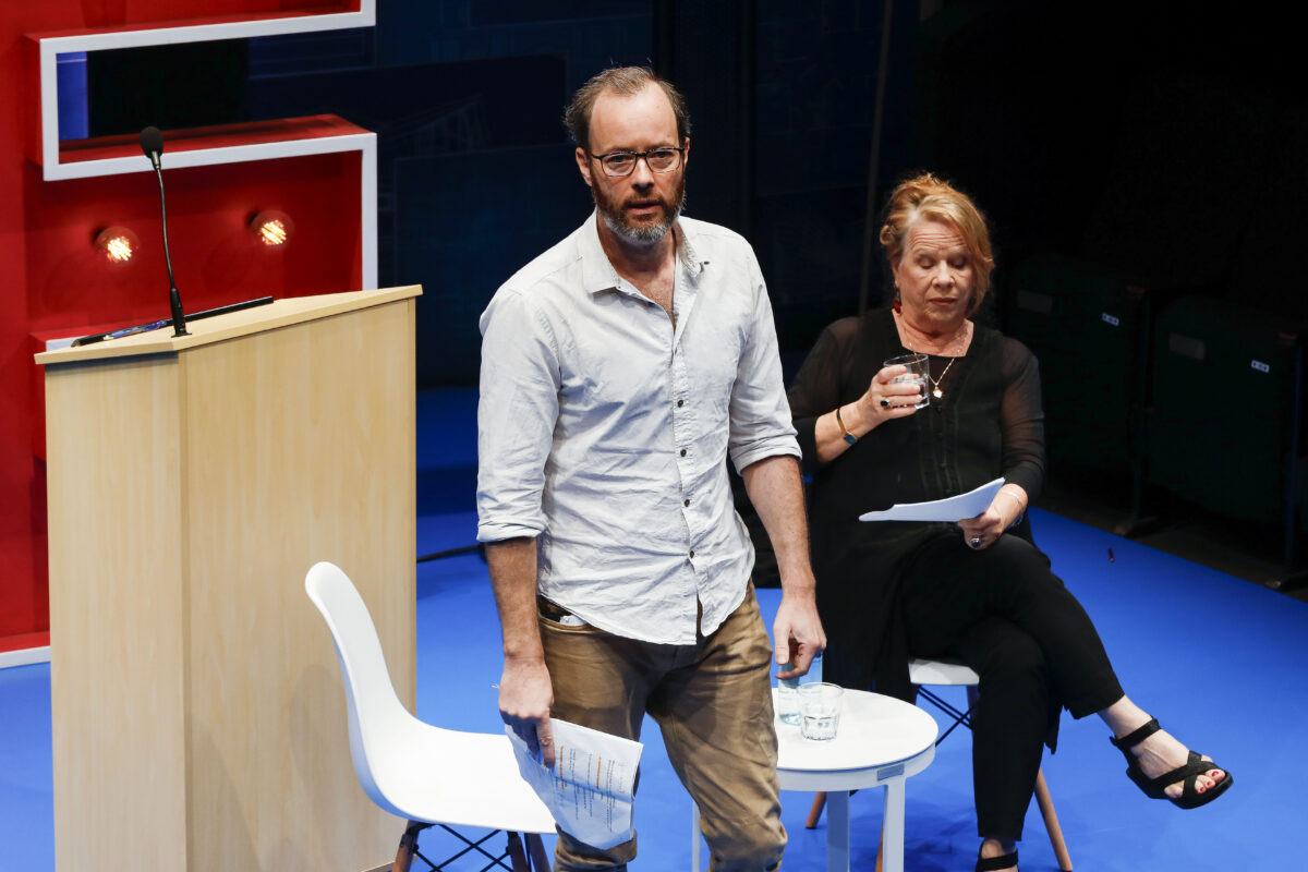 A man stands holding papers near a lectern, while a woman sits beside him with a glass and papers on her lap. Both appear to be on a stage during a presentation or discussion.